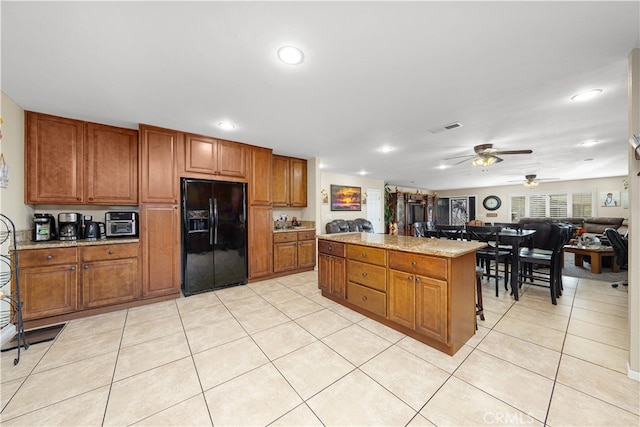 kitchen featuring ceiling fan, a kitchen island, black fridge with ice dispenser, light tile patterned flooring, and light stone counters