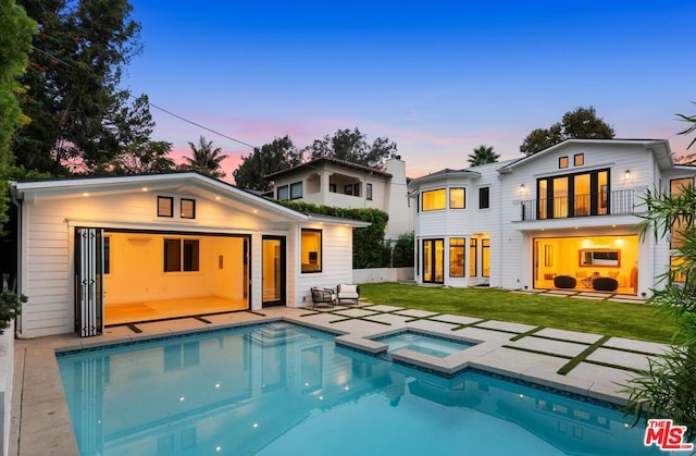 back house at dusk with a patio, a balcony, a lawn, and a pool with hot tub