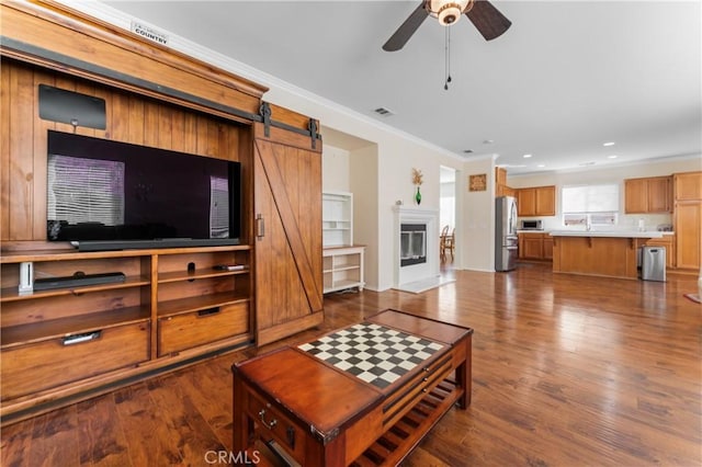 living room with ceiling fan, a barn door, dark wood-type flooring, and ornamental molding