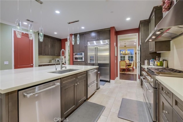 kitchen featuring a center island with sink, wall chimney range hood, sink, hanging light fixtures, and premium appliances