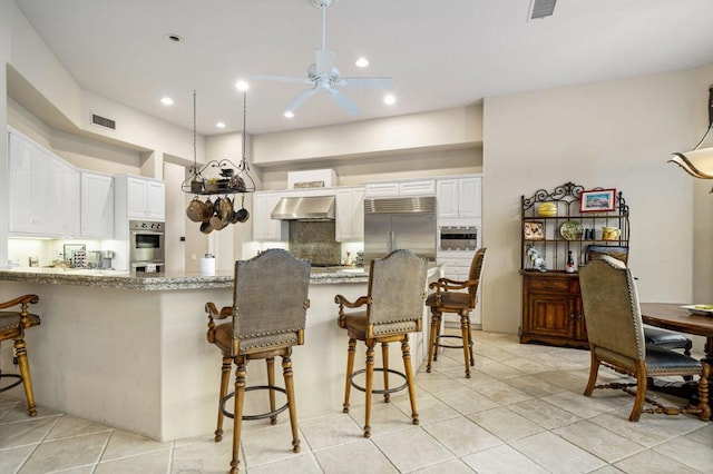 kitchen with light stone countertops, wall chimney exhaust hood, stainless steel appliances, ceiling fan, and white cabinets