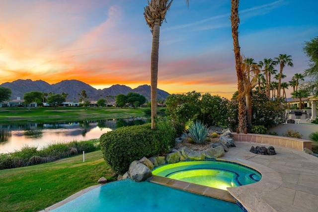 pool at dusk featuring an in ground hot tub, a water and mountain view, and a lawn