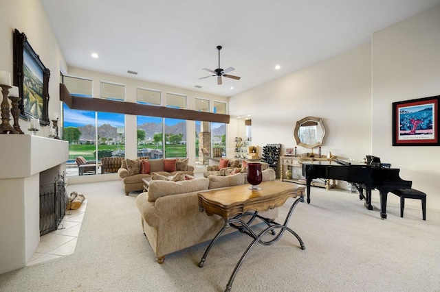 living room with ceiling fan, light colored carpet, a towering ceiling, and a tiled fireplace