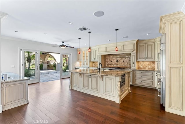 kitchen featuring an island with sink, decorative backsplash, light stone countertops, pendant lighting, and cream cabinets