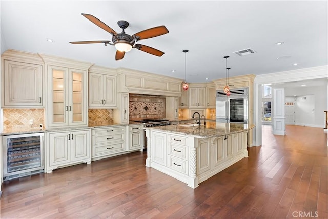 kitchen featuring pendant lighting, premium appliances, wine cooler, a kitchen island with sink, and cream cabinetry