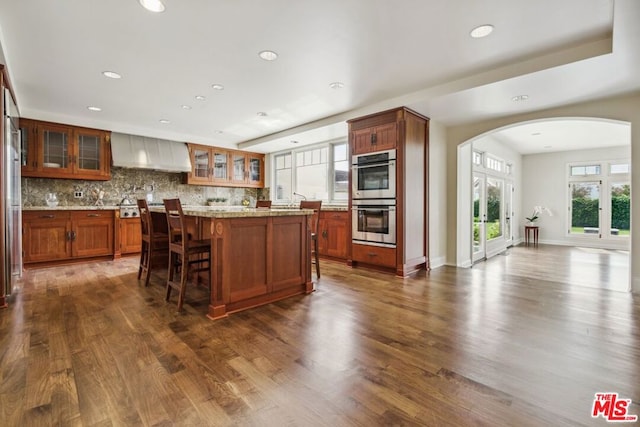 kitchen with backsplash, double oven, dark wood-type flooring, and wall chimney range hood