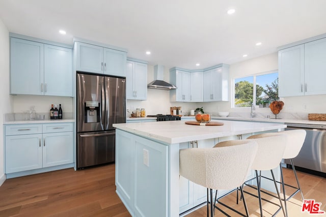 kitchen with wall chimney exhaust hood, a kitchen island, stainless steel appliances, a kitchen breakfast bar, and light hardwood / wood-style flooring