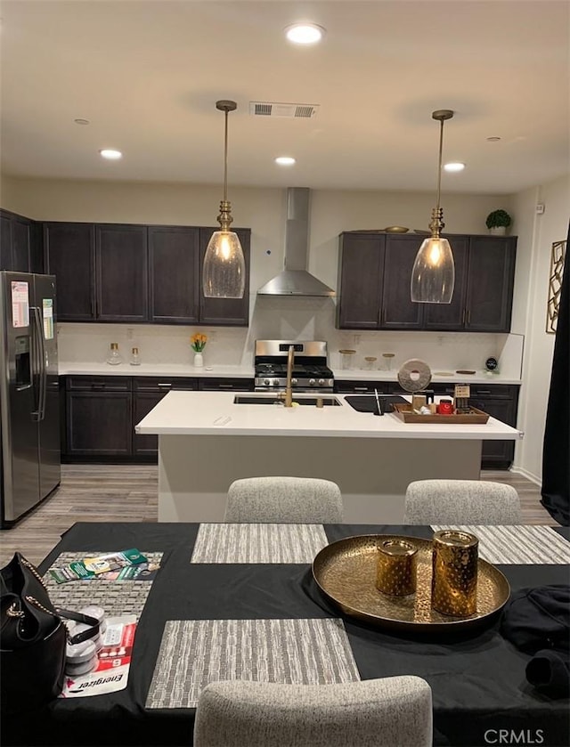 kitchen with appliances with stainless steel finishes, wall chimney range hood, pendant lighting, a center island with sink, and light hardwood / wood-style floors