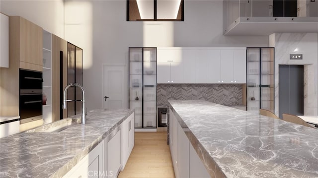 kitchen with black oven, light stone countertops, light wood-type flooring, white cabinets, and tasteful backsplash