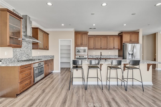 kitchen with a center island with sink, a breakfast bar area, light hardwood / wood-style floors, wall chimney exhaust hood, and stainless steel appliances