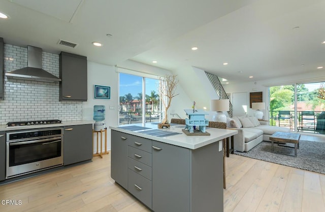 kitchen with wall chimney range hood, oven, light countertops, gray cabinets, and backsplash