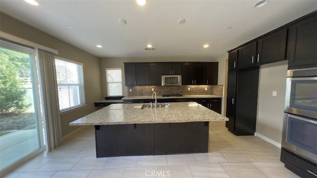 kitchen featuring sink, stainless steel appliances, light stone counters, and an island with sink