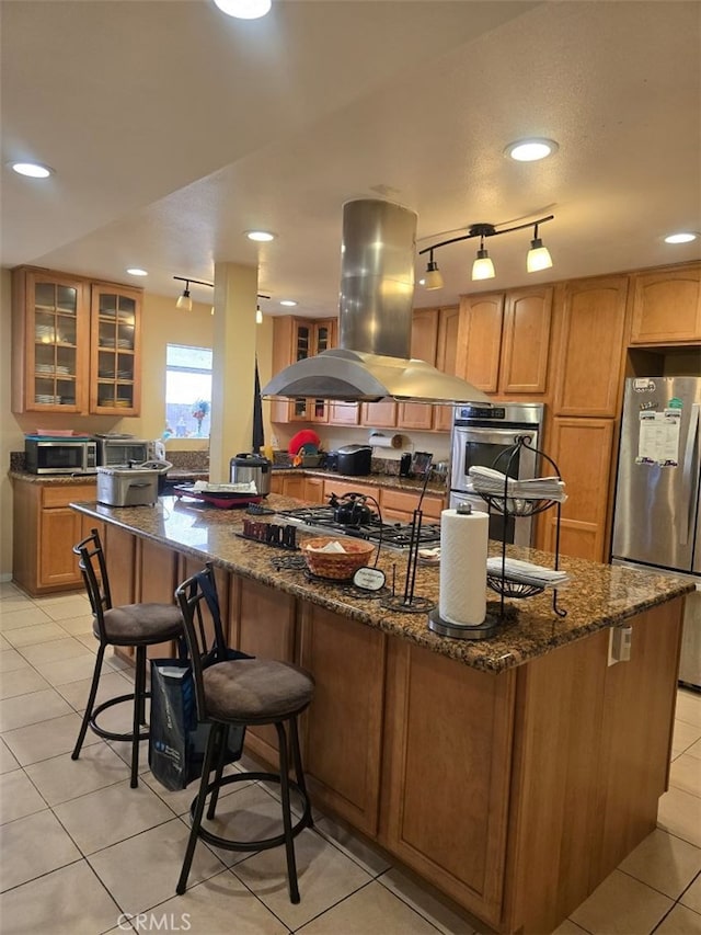 kitchen featuring island exhaust hood, appliances with stainless steel finishes, light tile patterned floors, and dark stone counters