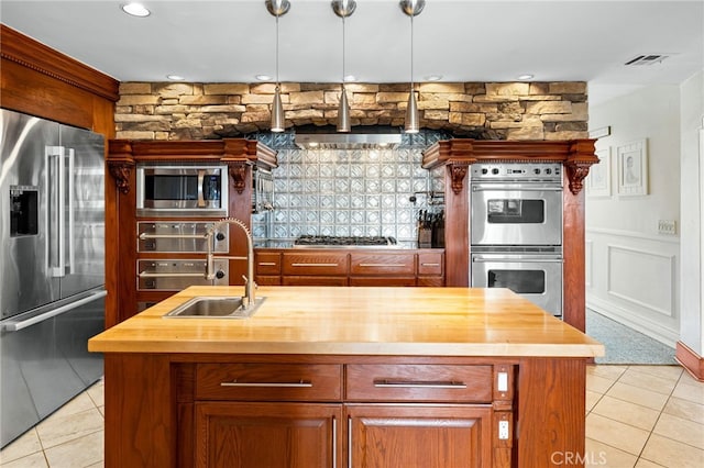 kitchen with wood counters, appliances with stainless steel finishes, a center island with sink, and hanging light fixtures