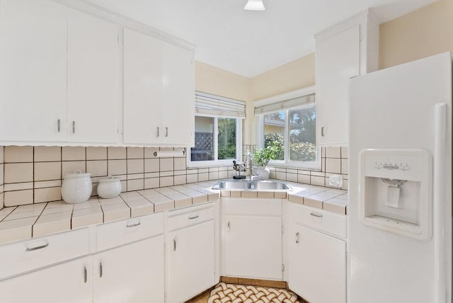 kitchen with tile countertops, white cabinetry, decorative backsplash, white refrigerator with ice dispenser, and sink