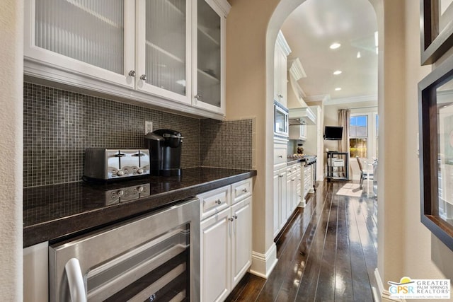 bar with dark wood-type flooring, crown molding, tasteful backsplash, beverage cooler, and white cabinets