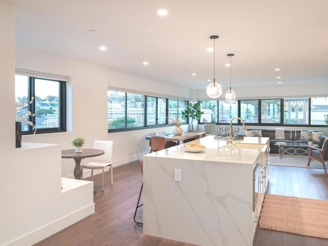 kitchen featuring decorative light fixtures, wood-type flooring, sink, light stone counters, and a center island with sink