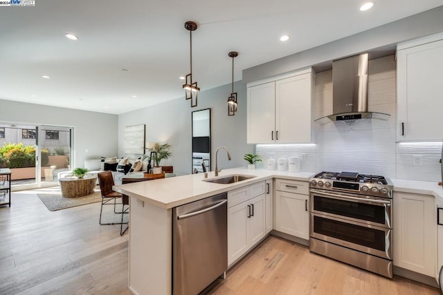 kitchen with appliances with stainless steel finishes, sink, light wood-type flooring, hanging light fixtures, and wall chimney exhaust hood