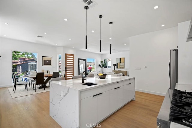 kitchen with a kitchen island with sink, white cabinets, sink, hanging light fixtures, and light hardwood / wood-style flooring