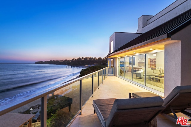 balcony at dusk featuring a view of the beach and a water view