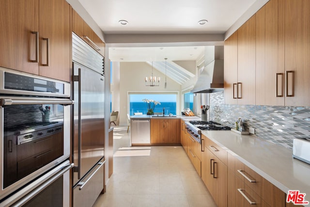 kitchen featuring wall chimney range hood, hanging light fixtures, stainless steel appliances, decorative backsplash, and an inviting chandelier