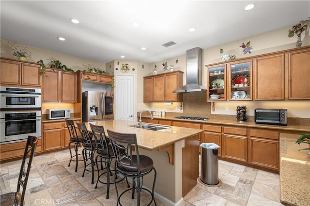 kitchen featuring wall chimney range hood, sink, appliances with stainless steel finishes, light stone counters, and a breakfast bar area