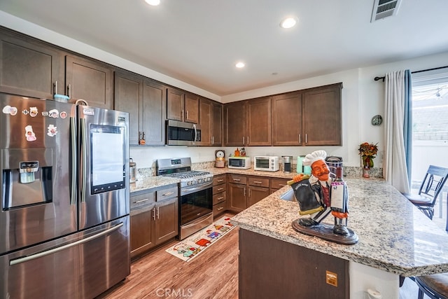 kitchen with kitchen peninsula, dark brown cabinets, appliances with stainless steel finishes, light hardwood / wood-style flooring, and light stone counters