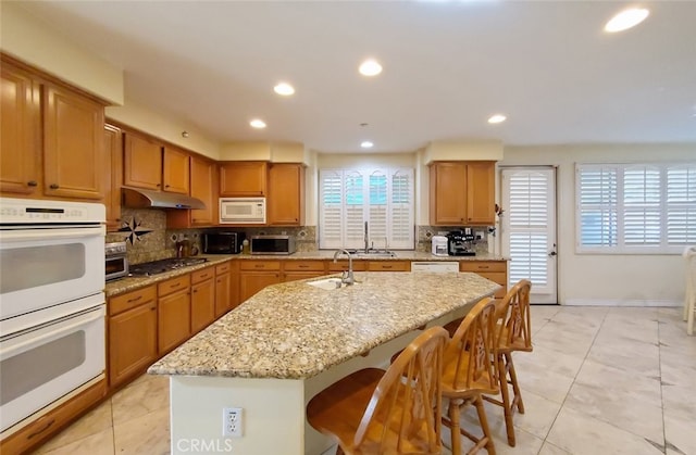 kitchen featuring white appliances, tasteful backsplash, light stone counters, under cabinet range hood, and a sink