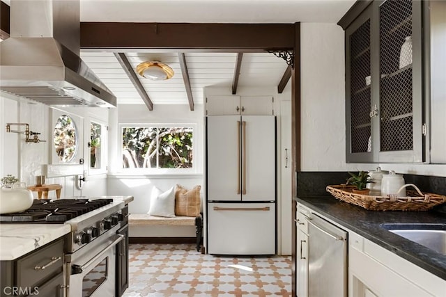 kitchen with premium appliances, wall chimney range hood, beam ceiling, dark stone countertops, and white cabinetry