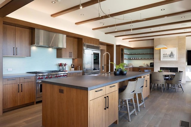 kitchen with a kitchen island with sink, wall chimney range hood, beamed ceiling, premium appliances, and dark hardwood / wood-style flooring