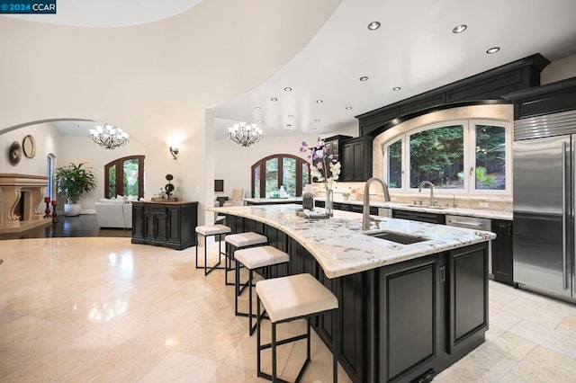 kitchen featuring french doors, a center island with sink, built in refrigerator, a notable chandelier, and light stone counters