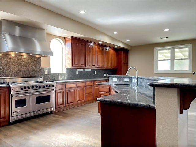 kitchen featuring range with two ovens, a center island with sink, tasteful backsplash, a sink, and island range hood