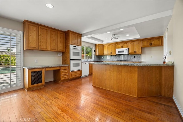 kitchen with kitchen peninsula, white appliances, a raised ceiling, ceiling fan, and light hardwood / wood-style floors