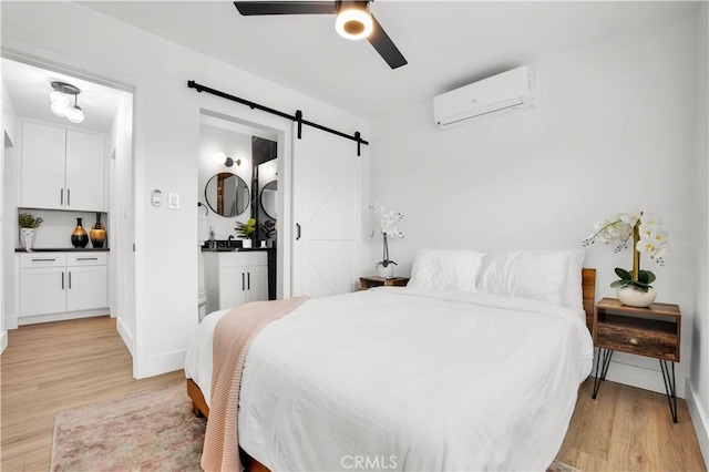 bedroom with ensuite bath, light hardwood / wood-style floors, a wall mounted AC, ceiling fan, and a barn door