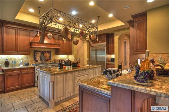 kitchen featuring high end appliances, a kitchen island with sink, dark stone counters, a raised ceiling, and a kitchen bar