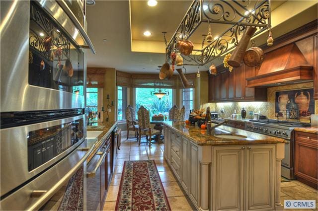 kitchen with sink, stainless steel appliances, dark stone counters, a center island with sink, and custom range hood