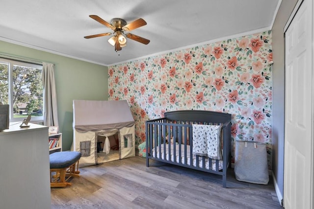 bedroom with hardwood / wood-style flooring, ornamental molding, a nursery area, and ceiling fan