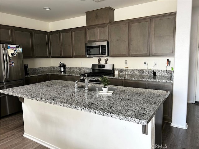 kitchen with a kitchen island with sink, dark hardwood / wood-style floors, light stone countertops, appliances with stainless steel finishes, and dark brown cabinets