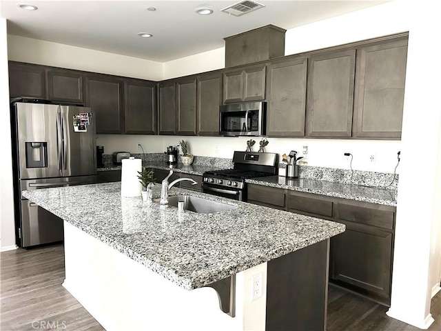 kitchen with light stone counters, dark brown cabinetry, stainless steel appliances, sink, and a center island with sink