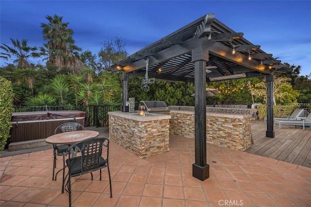 patio terrace at dusk featuring a grill, a hot tub, an outdoor kitchen, and a deck