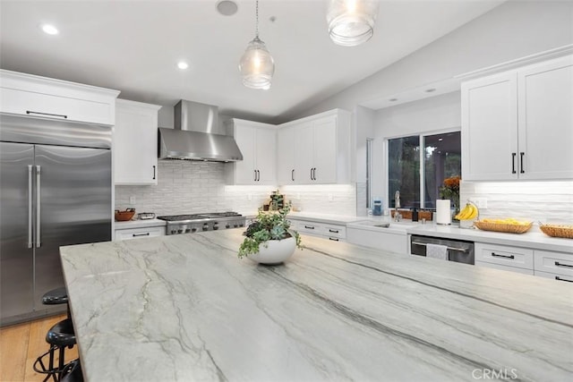 kitchen with pendant lighting, wall chimney exhaust hood, a kitchen bar, white cabinetry, and stainless steel appliances