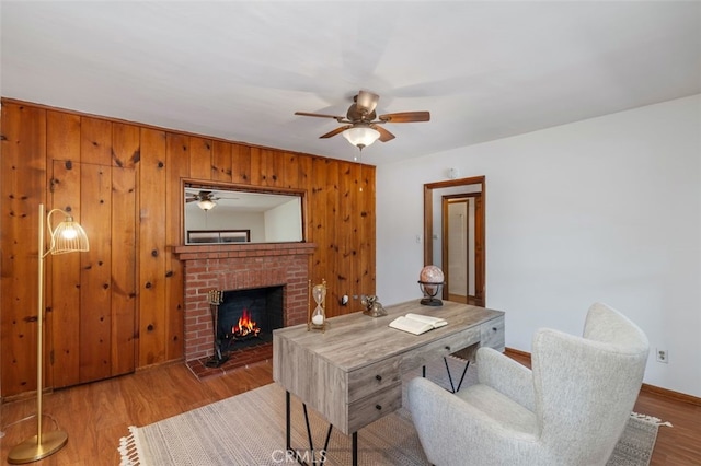 office area featuring ceiling fan, wood-type flooring, wooden walls, and a brick fireplace