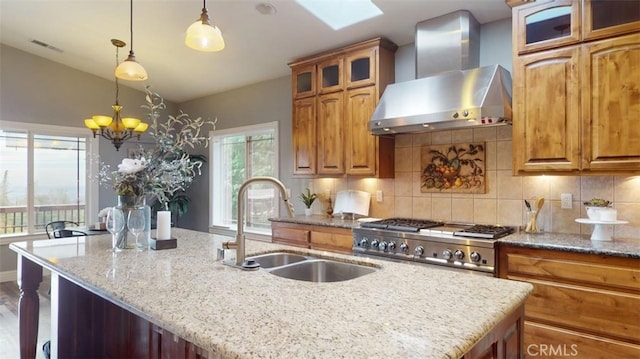 kitchen featuring hanging light fixtures, wall chimney exhaust hood, light stone counters, lofted ceiling with skylight, and a kitchen island with sink