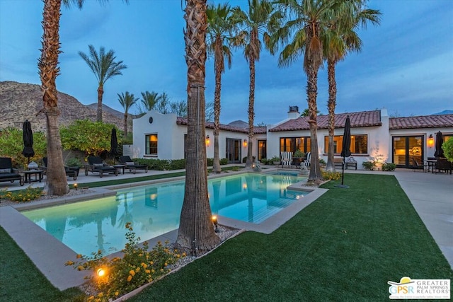 pool at dusk with a patio area, a mountain view, and a lawn