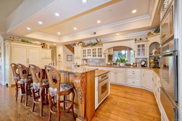 kitchen with a kitchen island with sink, a raised ceiling, light stone counters, light hardwood / wood-style floors, and decorative backsplash