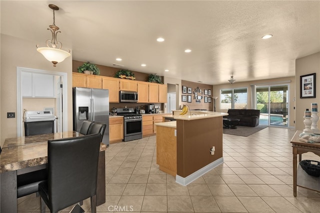 kitchen featuring appliances with stainless steel finishes, light brown cabinets, light tile patterned floors, decorative light fixtures, and an island with sink