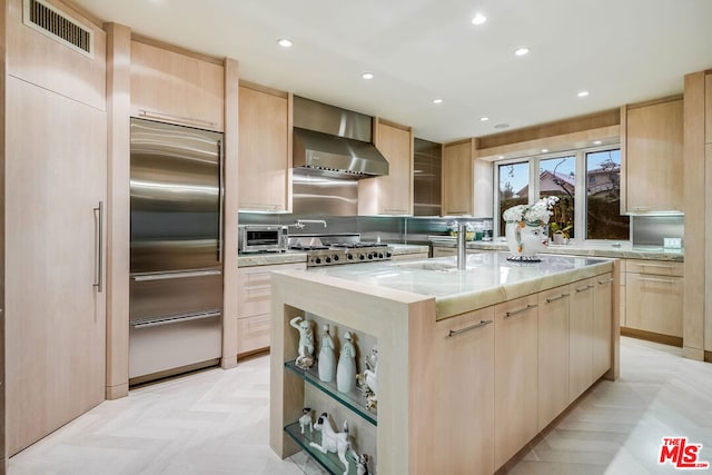 kitchen featuring stainless steel appliances, light parquet floors, wall chimney range hood, light brown cabinets, and an island with sink