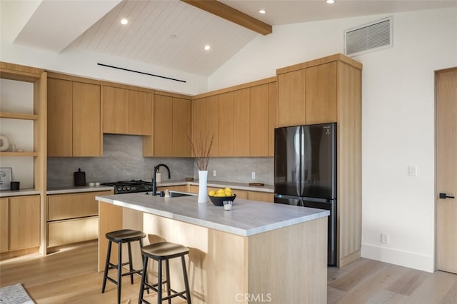 kitchen featuring tasteful backsplash, black fridge, light hardwood / wood-style floors, beamed ceiling, and a center island with sink