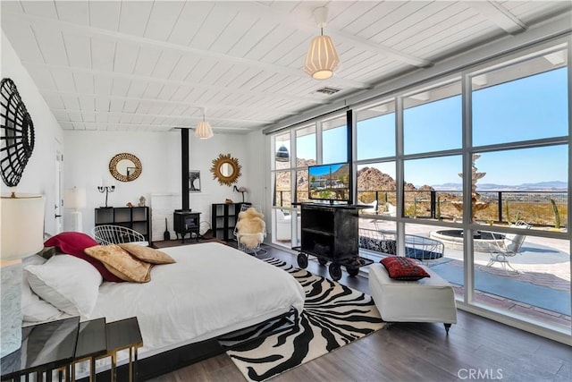 bedroom featuring a wood stove, a wall of windows, wood finished floors, and beamed ceiling