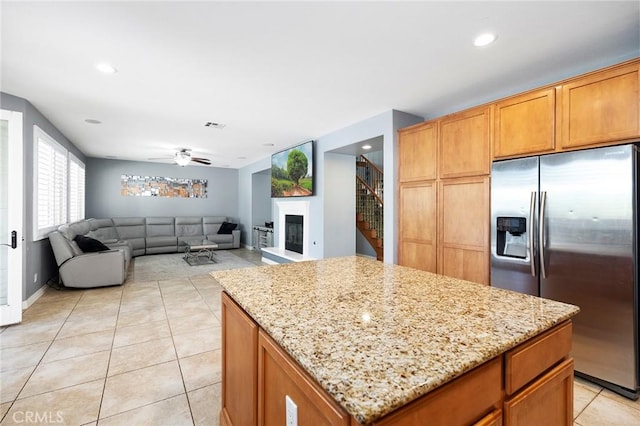 kitchen with ceiling fan, a center island, stainless steel fridge with ice dispenser, light tile patterned floors, and light stone counters